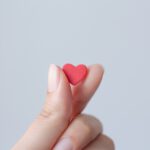 Eine manikürte Frauenhand hält ein kleines rotes Herz aus Papier zwischen Daumen und Zeigefinger.