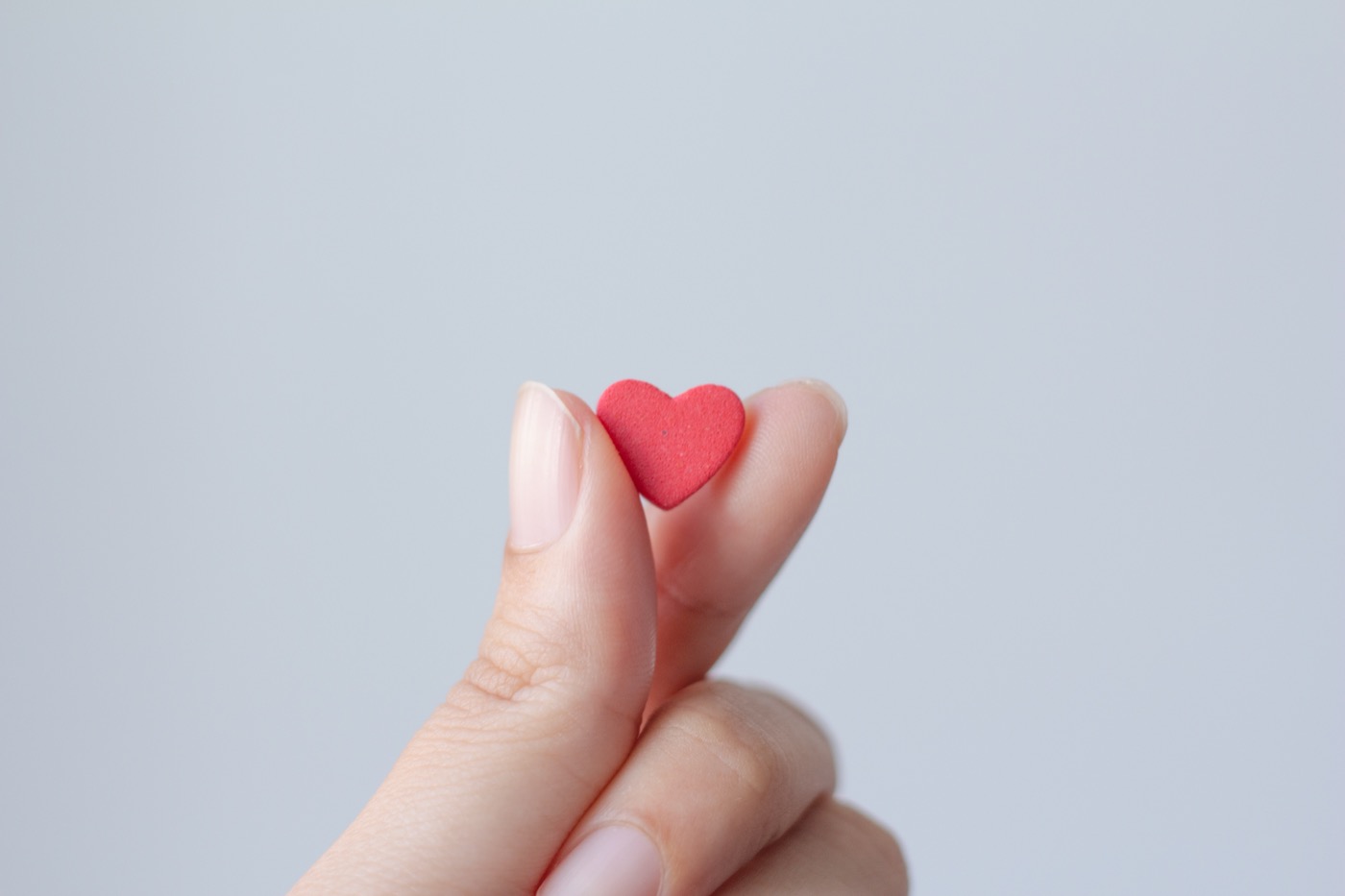 Eine manikürte Frauenhand hält ein kleines rotes Herz aus Papier zwischen Daumen und Zeigefinger.
