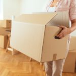 Eine Person trägt einen großen, geöffneten Umzugskarton in einer Wohnung mit weiteren gepackten Kartons im Hintergrund.