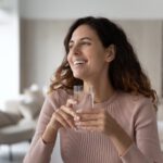 Eine lächelnde Frau sitzt in einem hellen Wohnzimmer und hält ein Glas klares Wasser in beiden Händen.