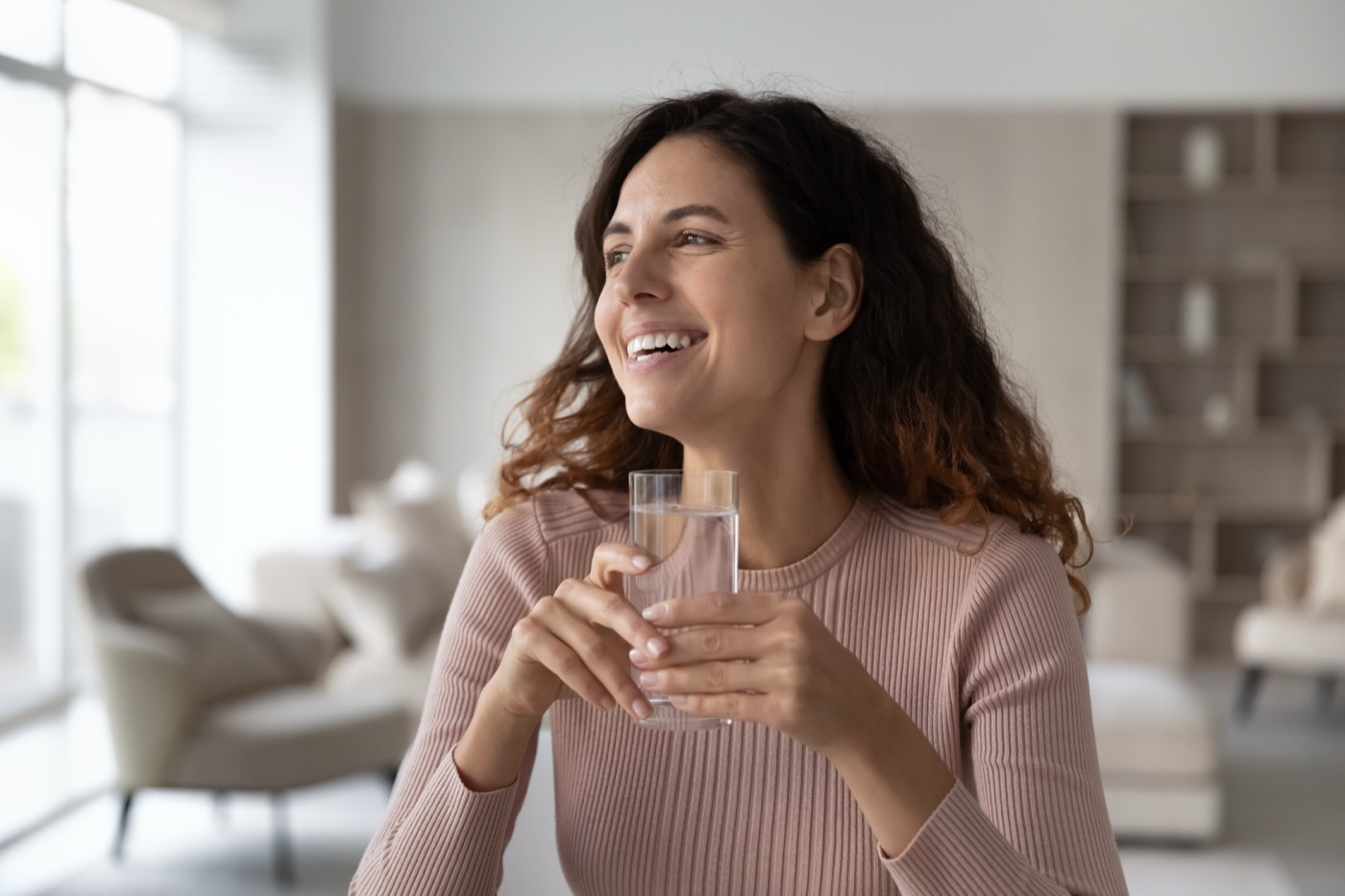 Eine lächelnde Frau sitzt in einem hellen Wohnzimmer und hält ein Glas klares Wasser in beiden Händen.