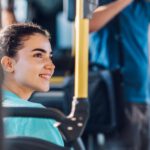 Eine lächelnde junge Frau fährt im Stadtbus und blickt aus dem Fenster, während im Hintergrund ein anderer Fahrgast steht.