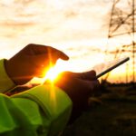 Nahaufnahme der Hände einer Person in Warnkleidung, die bei Sonnenuntergang vor einem Strommast ein Tablet bedient.