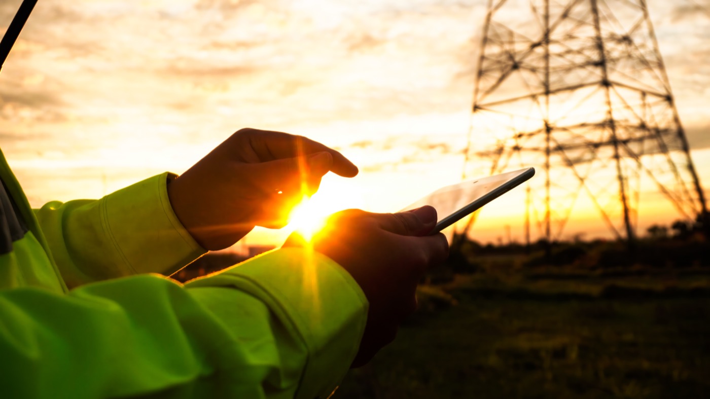 Nahaufnahme der Hände einer Person in Warnkleidung, die bei Sonnenuntergang vor einem Strommast ein Tablet bedient.