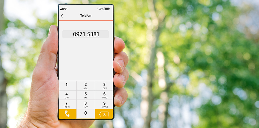 Eine Hand hält ein Smartphone, auf dessen Bildschirm die Telefonnummer 0971 5381 eingegeben ist, vor einem unscharfen grünen Hintergrund.