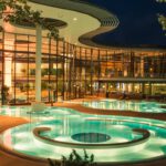 Die beleuchteten Außenbecken der KissSalis Therme bei Nacht vor der gläsernen, modernen Architektur des Hauptgebäudes.