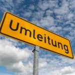 Ein gelbes Verkehrsschild mit der Aufschrift „Umleitung“ an einem Metallpfosten vor einem leicht bewölkten, blauen Himmel.