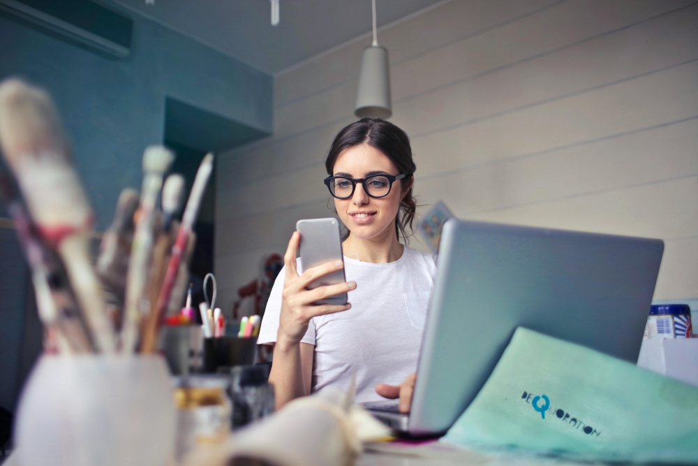 Eine lächelnde Frau mit Brille blickt auf ihr Smartphone, während sie an einem Schreibtisch vor einem Laptop sitzt.