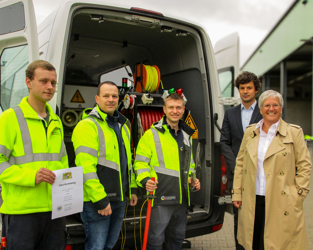 Drei Techniker in Warnkleidung und zwei Führungskräfte posieren lächelnd vor einem geöffneten Service-Transporter, während einer eine Anerkennungsurkunde hält.