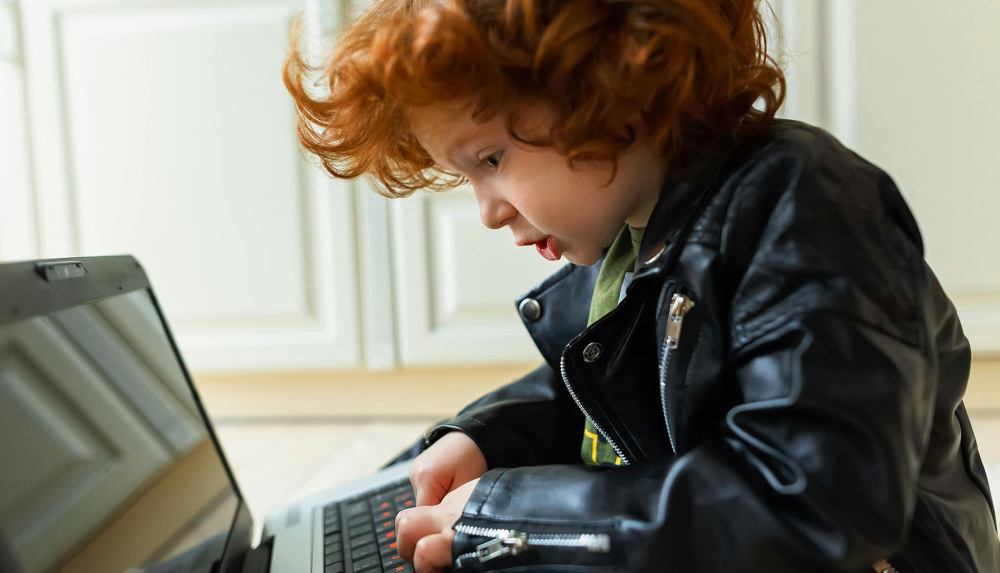Kinderleichte Online-Services: Ein Kind mit lockigen roten Haaren und Lederjacke blickt konzentriert auf die Tastatur eines geöffneten Laptops.