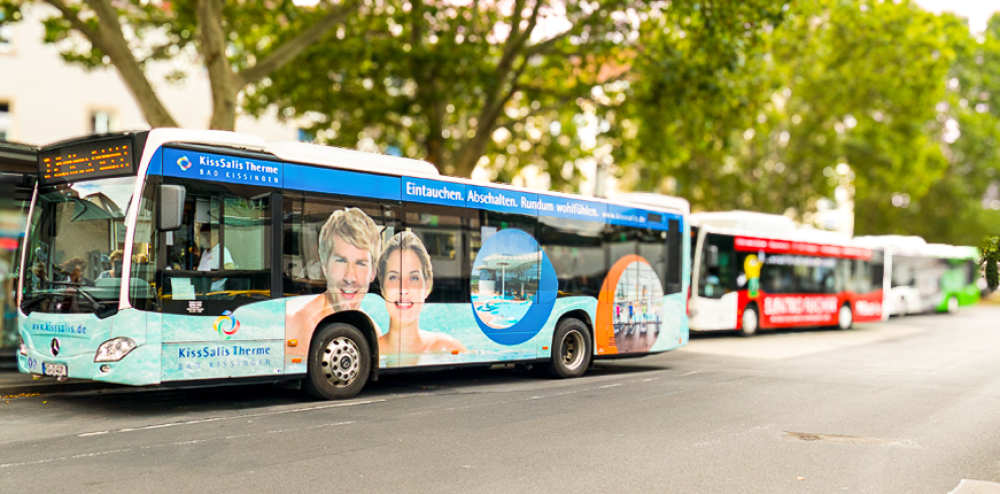Ein Stadtbus mit einer großflächigen Werbeaufschrift der KissSalis Therme steht an einer Haltestelle in einer Reihe mit weiteren Bussen.