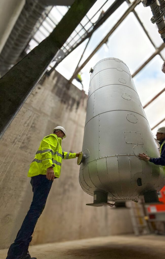 Zwei Techniker in einer Heizzentrale begleiten die Installation eines großen, zylindrischen Metalltanks, der an einem Kran hängt.