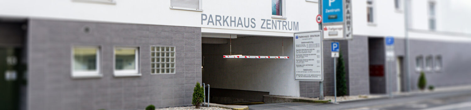 Die Einfahrt des Parkhauses Zentrum mit einer Schrankenanlage und einer Infotafel zu Tarifen rechts neben der Zufahrt.