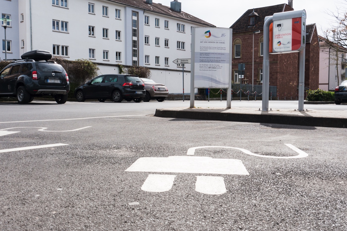Ein markierter Parkplatz für Elektroautos mit einem großen, aufgemalten Steckersymbol auf dem Asphalt vor einer E-Ladestation.
